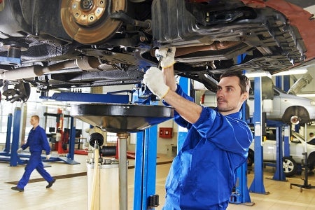 A mechanic in blue overalls repairing the undercarriage of a raised car.